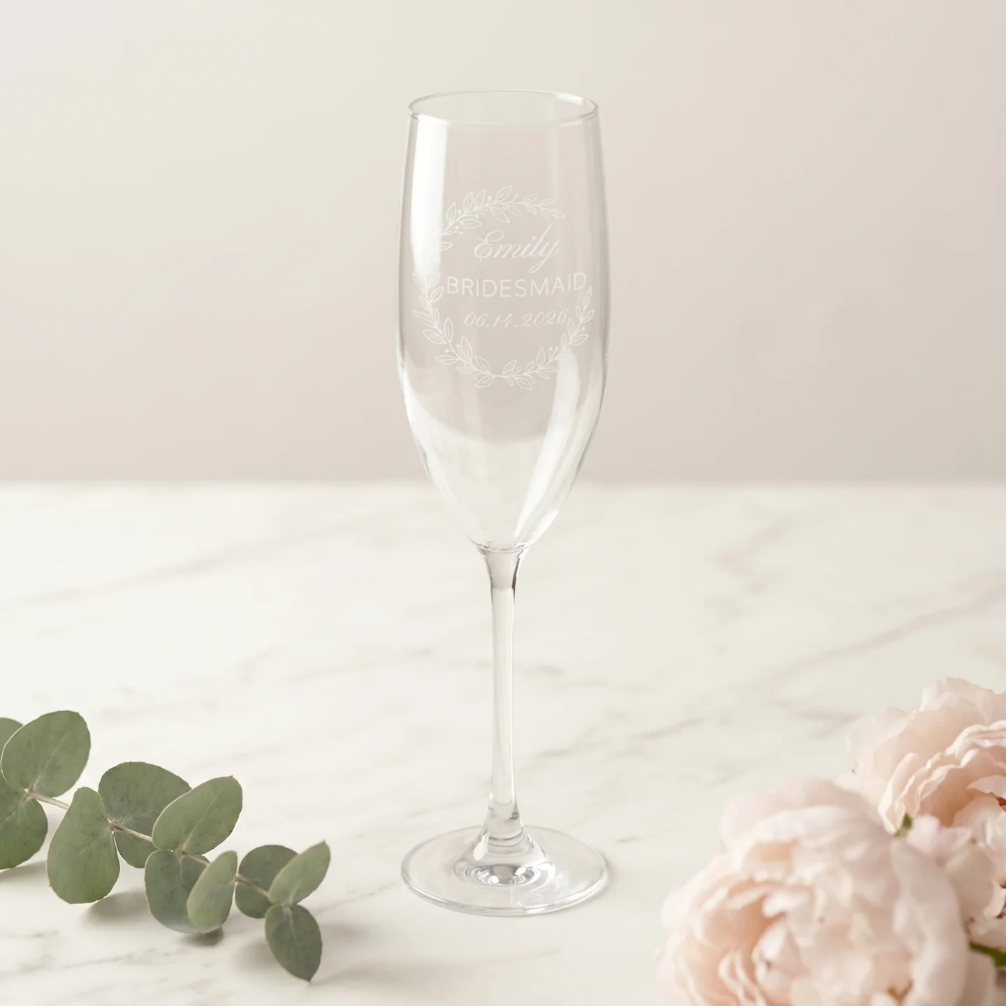 A bridesmaid proposal gift — a personalized champagne flute with the Eucalyptus Crescent Crest design, a soft eucalyptus arch above a cursive first name, the word 'Bridesmaid' in small caps, and a wedding date — on a white marble counter.