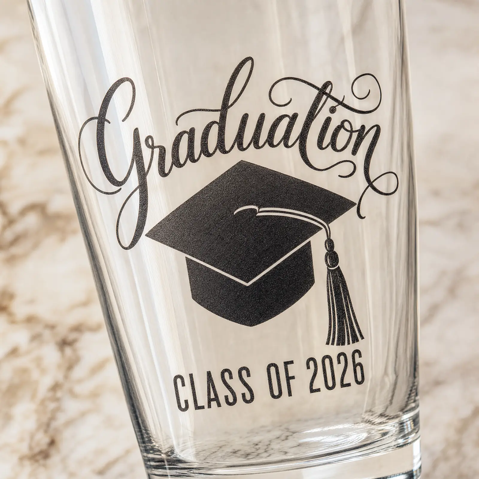 Macro closeup of the engraving — cursive 'Graduation' on top, black graduation cap silhouette in the middle, 'Class of 2026' in condensed sans-serif beneath.