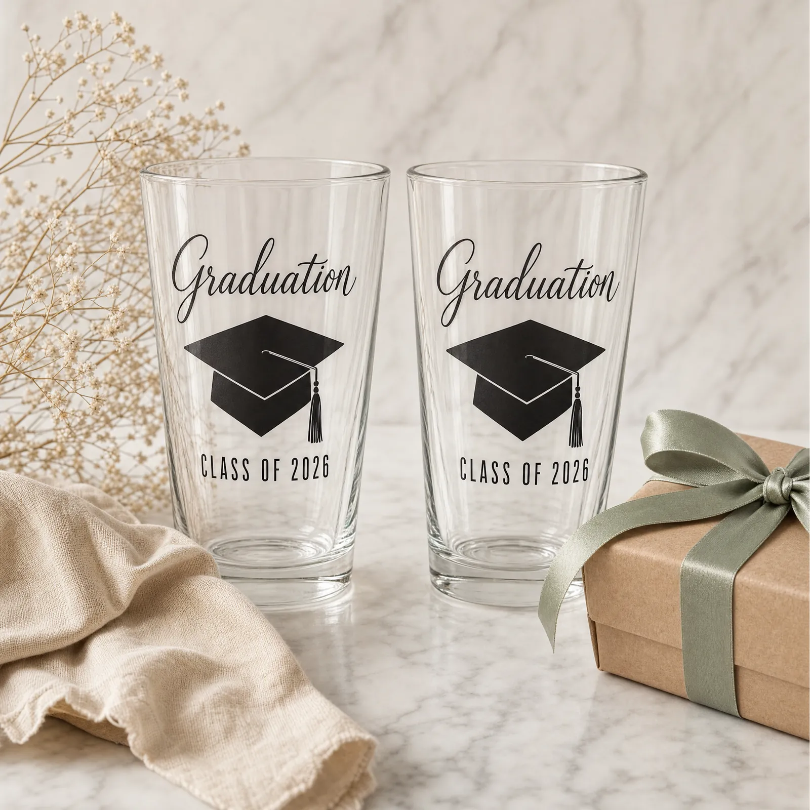 Two matching personalized pint glasses standing side by side on white marble with a kraft gift box tied with sage-green satin ribbon and a sprig of dried baby's breath.
