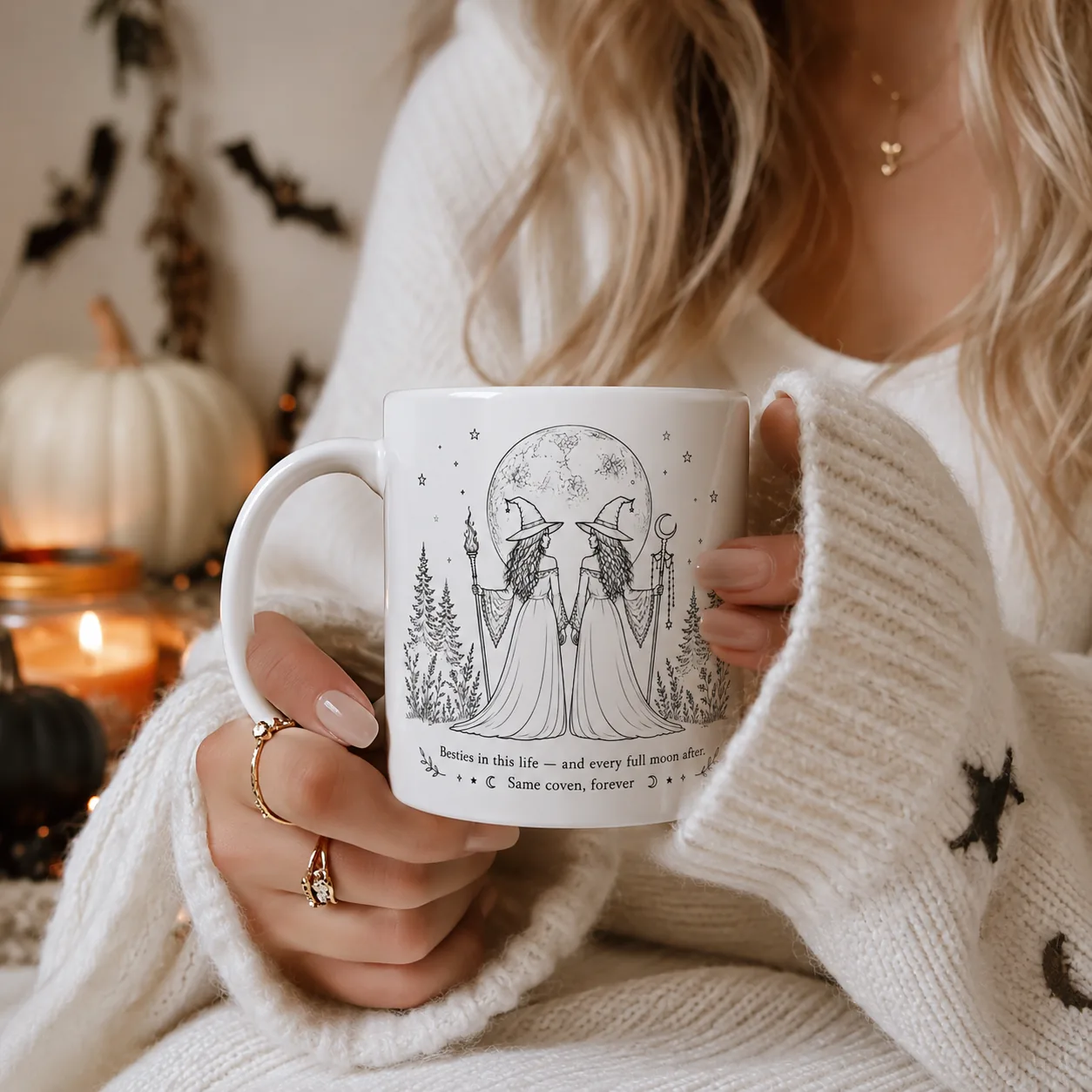 Woman in a cream knit sweater holding a personalized besties witch mug — two witches holding hands under a full moon with pine trees on either side, printed with the quote Besties in this life, and every full moon after, same coven forever, styled with candlelight and white pumpkins.