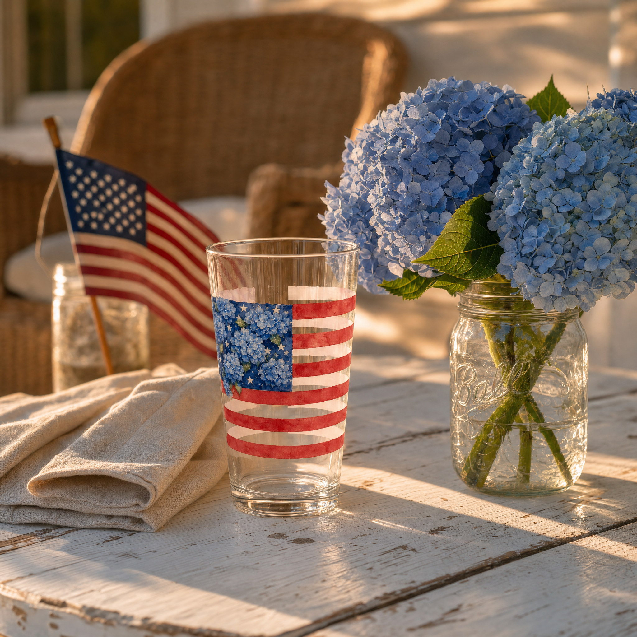 A softly styled porch table with cream linen, a personalized Hydrangea Flag pint glass, blue hydrangeas in a jar, and soft afternoon light — no faces.