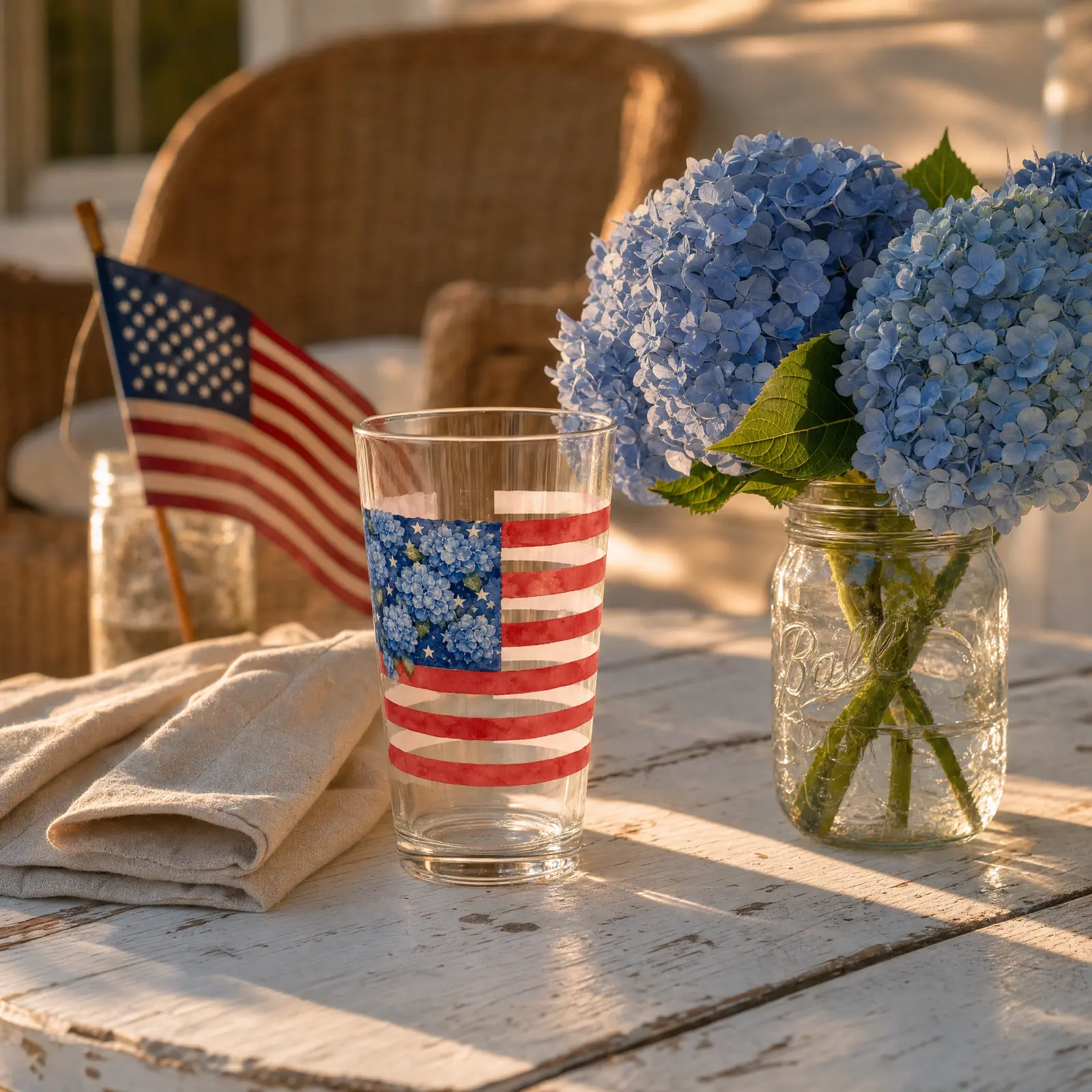 A softly styled porch table with cream linen, a personalized Hydrangea Flag pint glass, blue hydrangeas in a jar, and soft afternoon light — no faces.