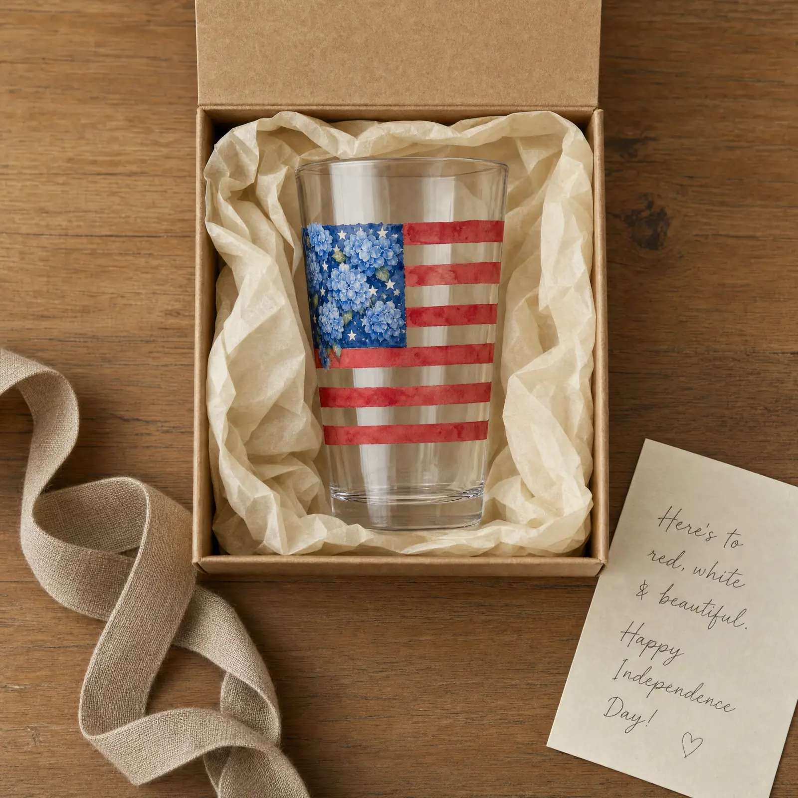 A personalized Hydrangea Flag pint glass in a kraft gift box with cream tissue and a sage ribbon, on white marble, soft daylight.