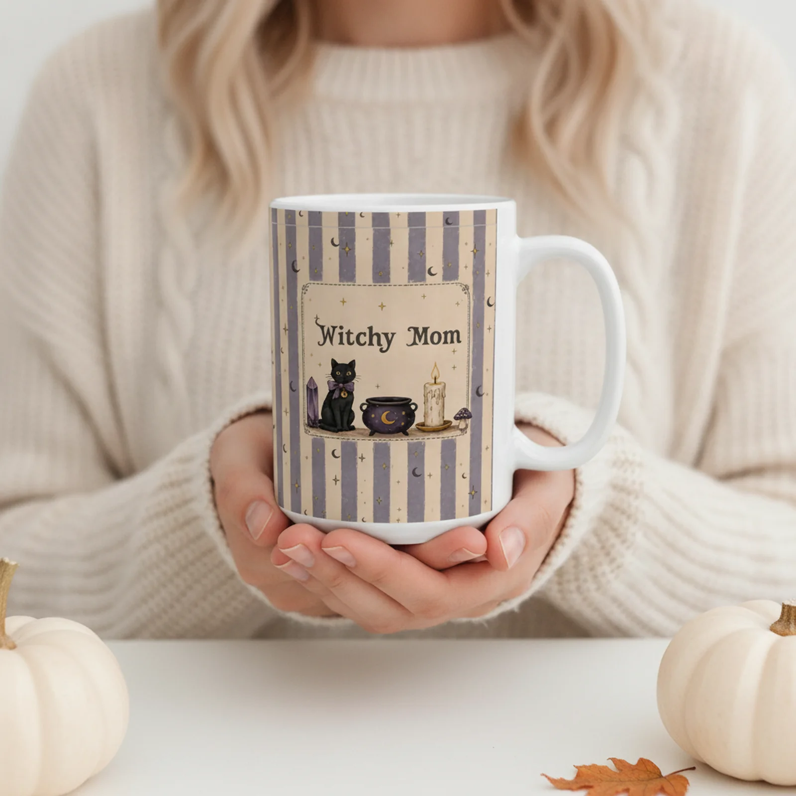 Woman in a cream knit sweater holding a personalized Witchy Mom mug with a black cat, cauldron and candle design in soft purple stripes, white pumpkins nearby.